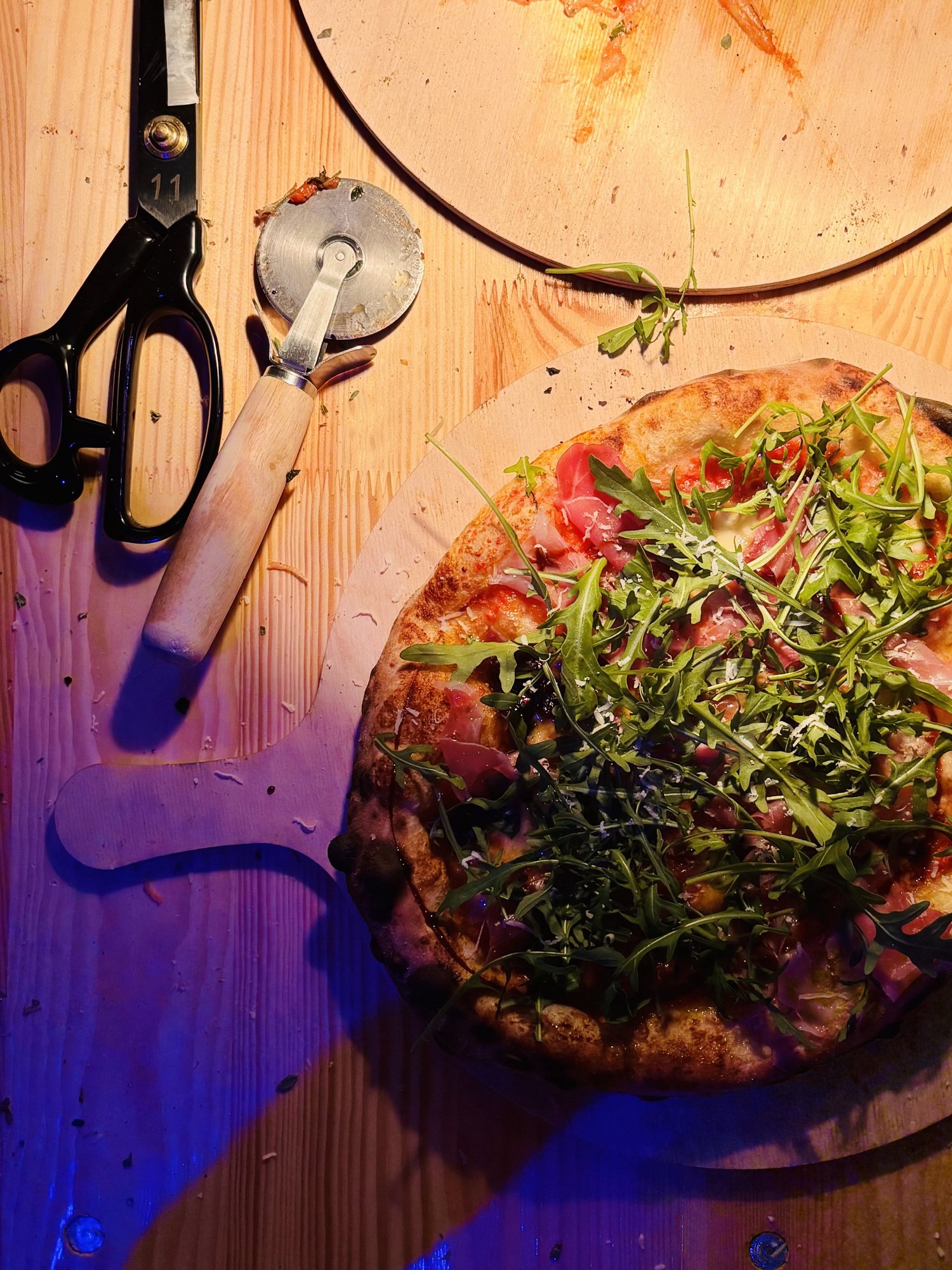 Fertige Pizza mit Rucola und Parmesan auf dem Holzbrett, daneben Pizzaroller und Schere